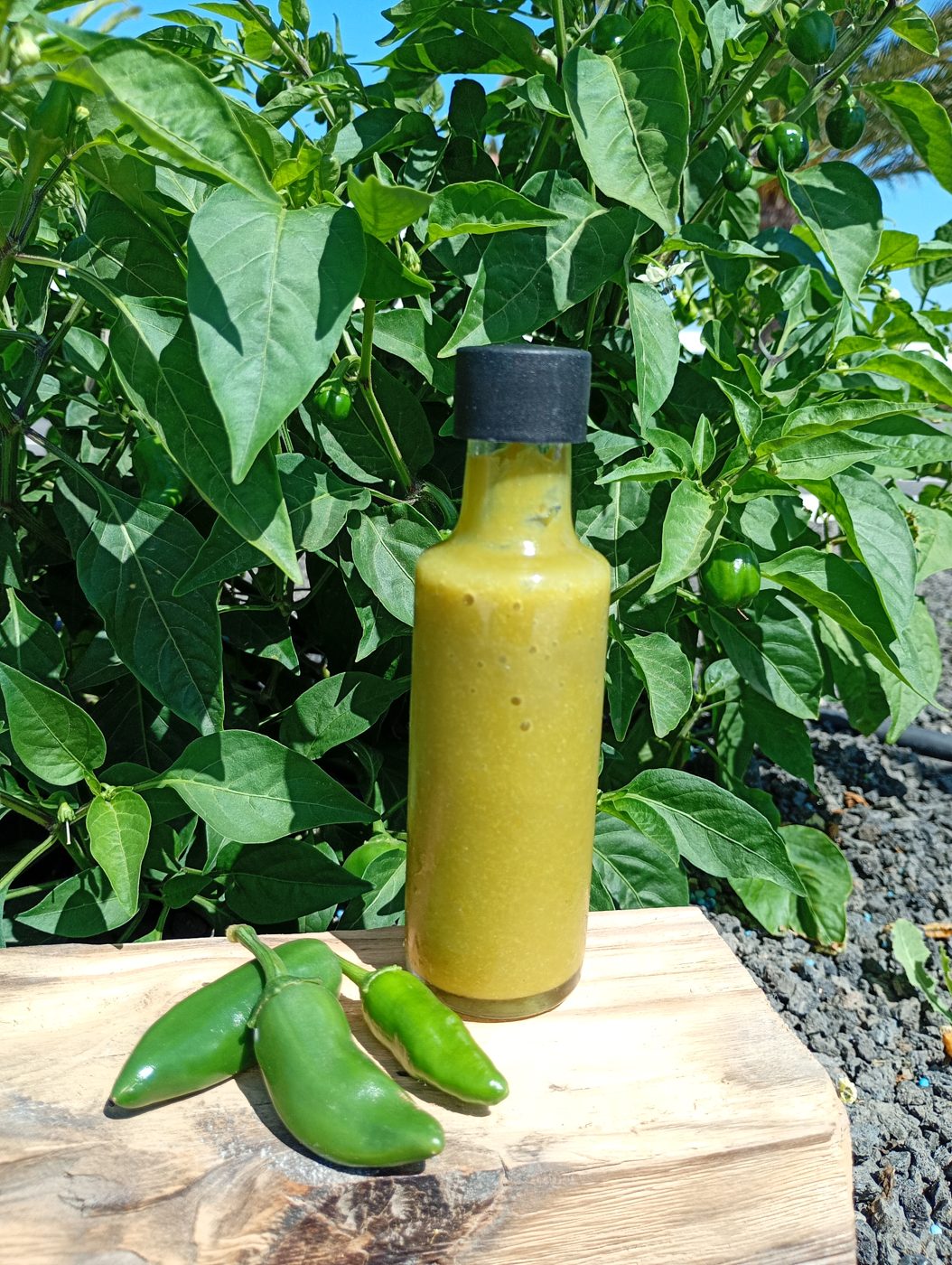 Botella de salsa picante verde casera entre matas de chile y unos chiles verdes sobre una tabla, en Lajares
