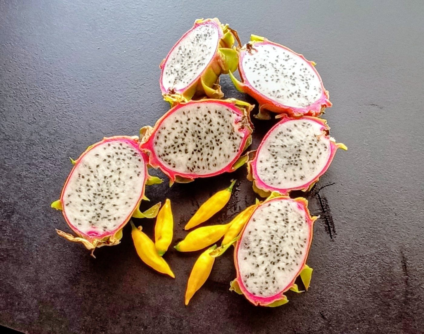 Pitahayas (fruta del dragón) cortadas por la mitad sobre una mesa oscura, con chiles amarillos alrededor