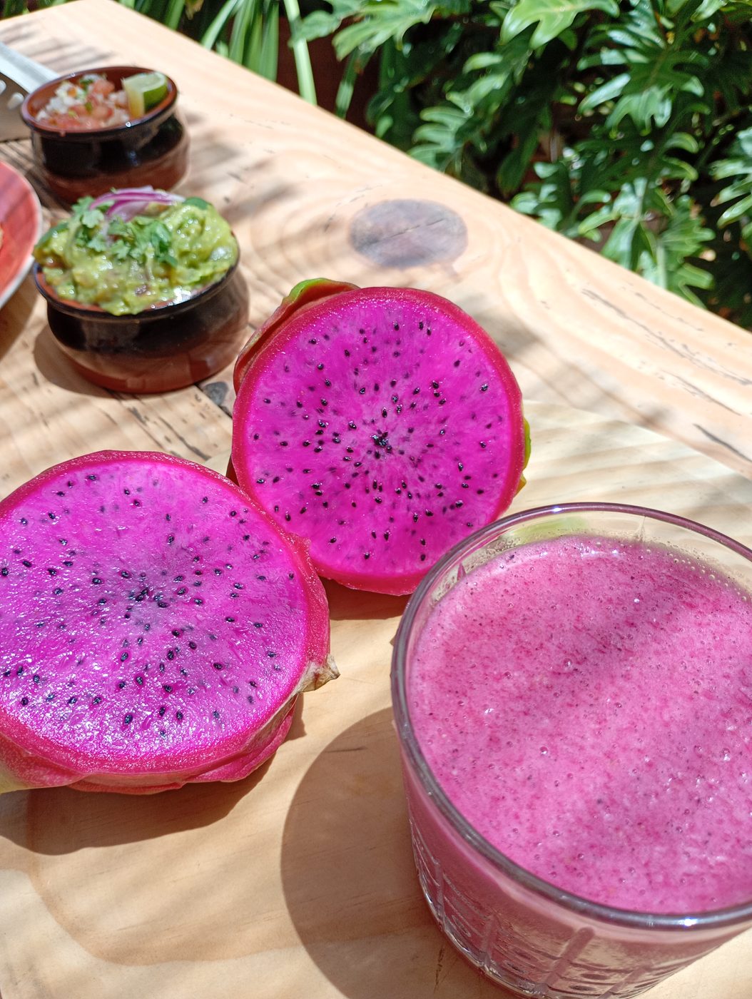 Vaso de smoothie Tropicalia de color rosa intenso junto a pitahayas cortadas, sobre una mesa de la terraza